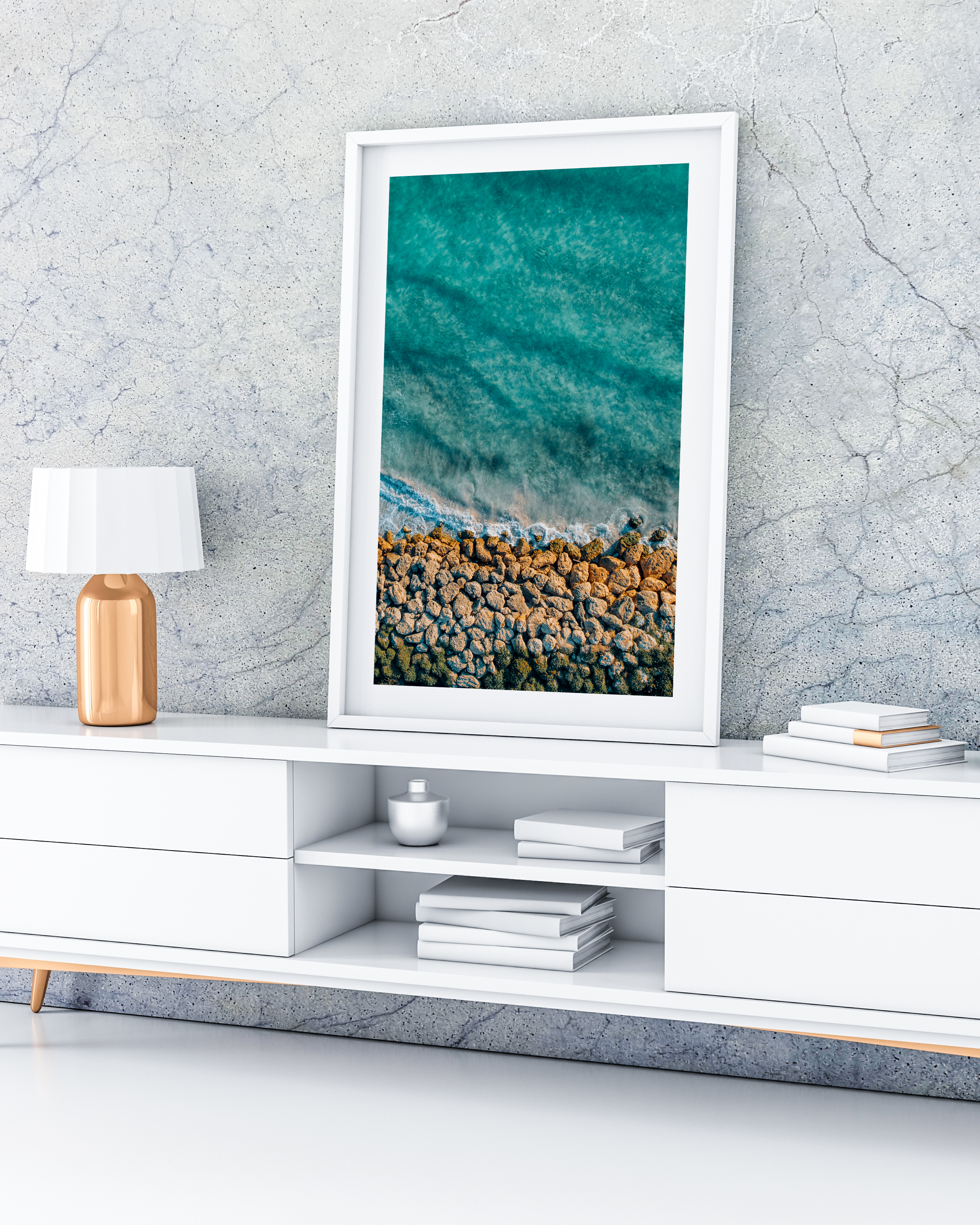 A print of a rocky shoreline in a white frame, sitting on a white buffet. There is a grey wall behind it and a white and gold lamp next to it.